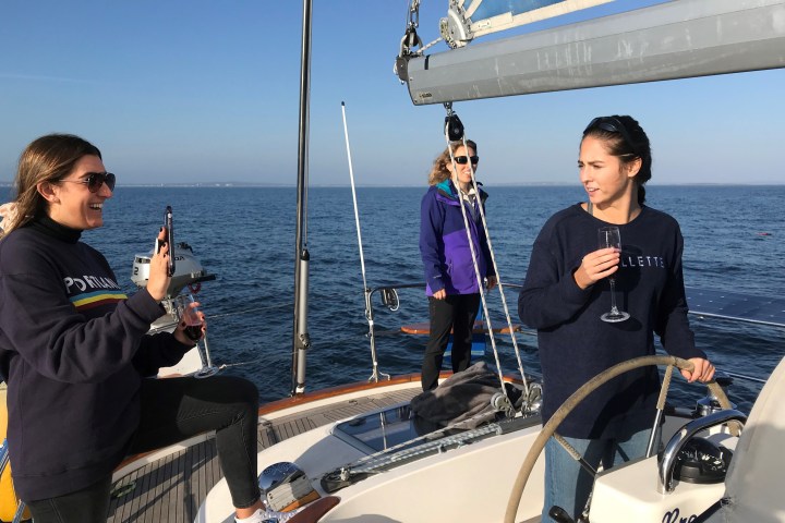 Three women on a sailboat, one steering, another taking a photo, and the third standing, all in casual attire.