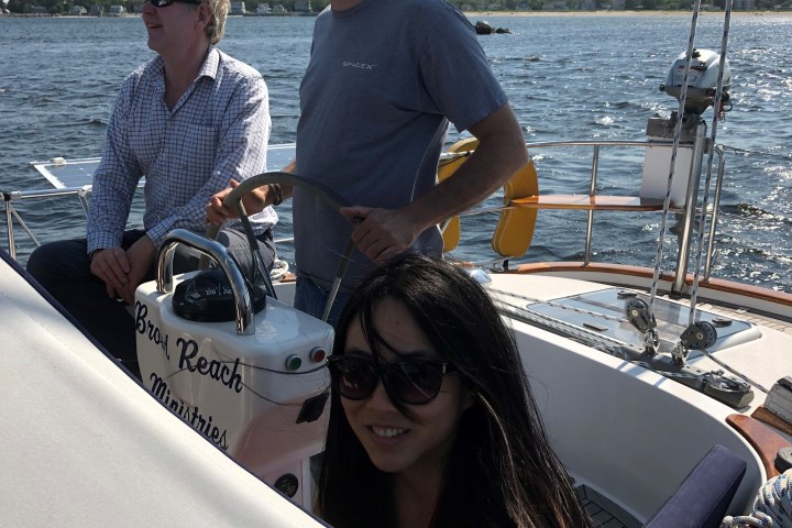 Three people enjoying a sunny day on a sailboat, with two standing and one sitting at the helm.