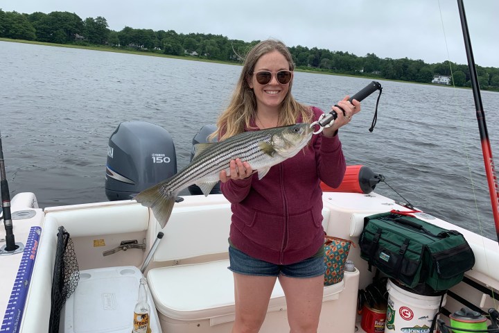 Woman on boat holding a striped bass fish, smiling by a lake.