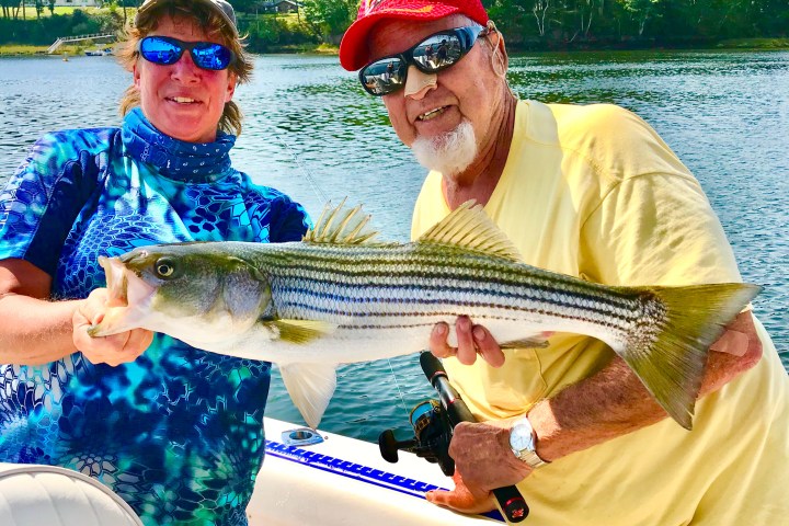 Two people hold a large fish on a boat with water and trees in the background.