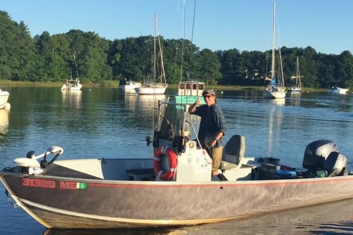 Man on small motorboat waves on a calm lake with sailboats and trees nearby.