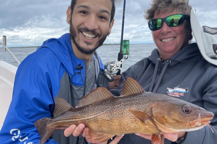 Two people smiling, holding a large fish on a boat with a cloudy sky.