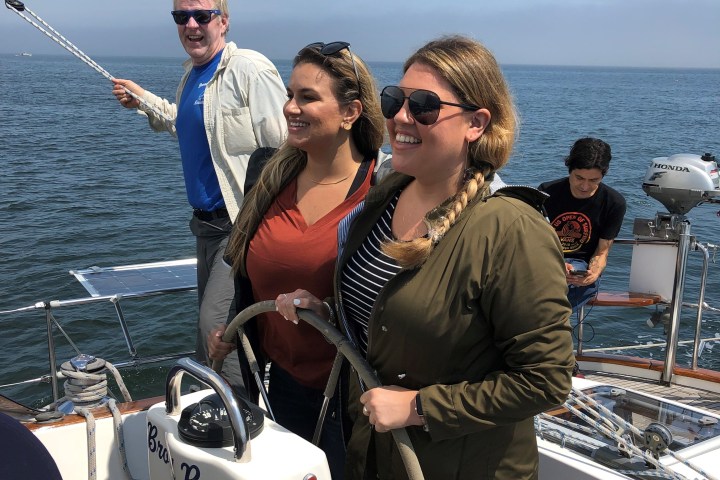 Three people smiling on a sailboat at sea, with a man holding a rope and two women at the helm.
