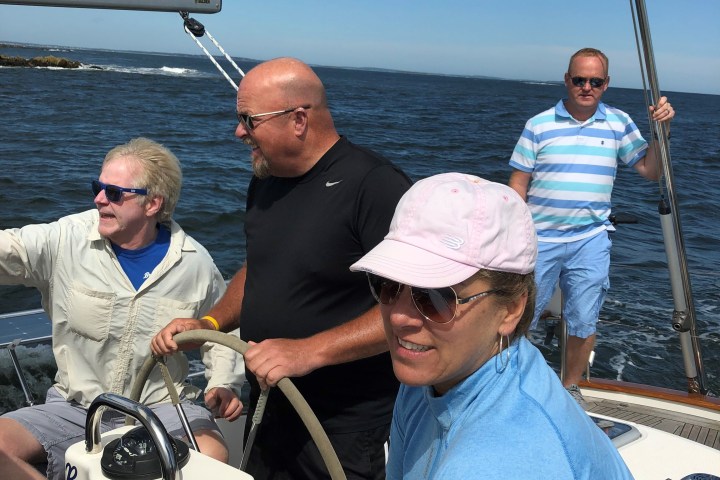 Four people sailing on a boat with a man steering and others enjoying the ocean view under sunny skies.