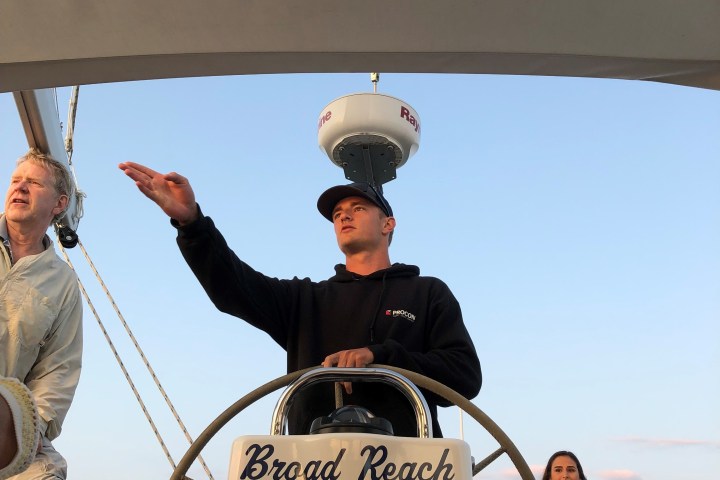 Three people on a sailboat named 'Broad Reach', one steering and pointing, with a clear sky background.