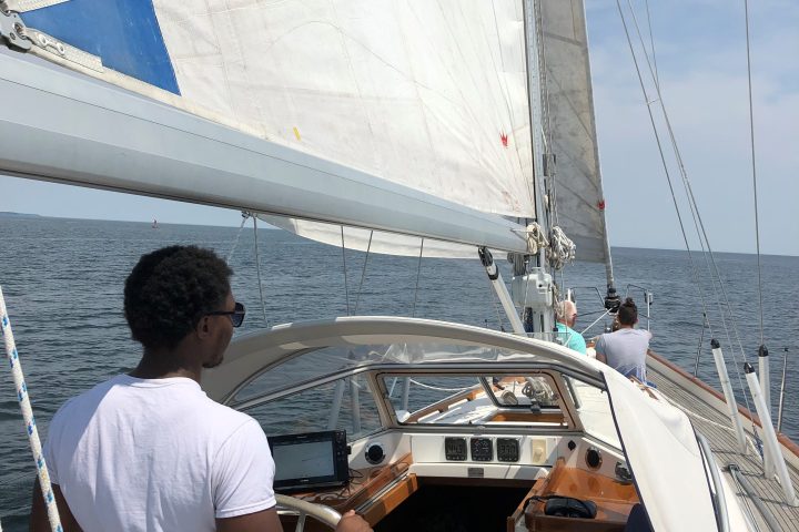 Person steering a sailboat with a large white sail on a sunny day, ocean in the background.