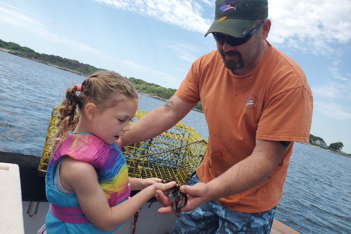 Child and adult on a boat holding a small sea creature with a lobster trap in background.