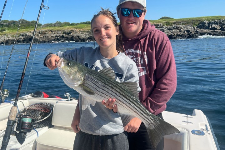 Two people on a boat holding a large fish, with fishing rods and water in the background.