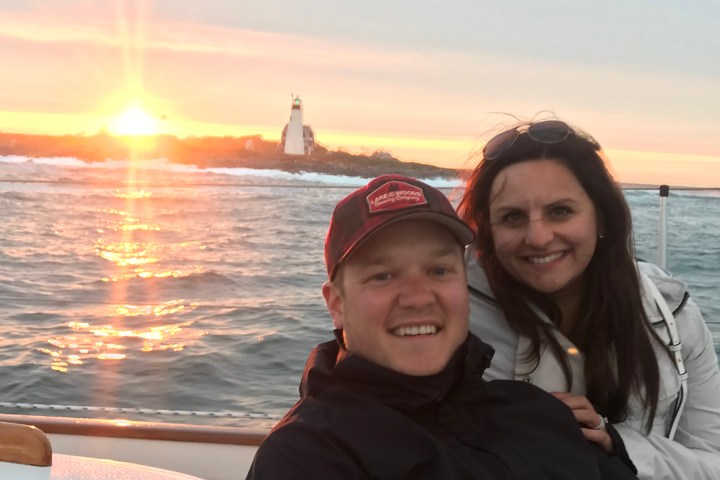 Two people smiling on a boat at sunset with a lighthouse in the background.