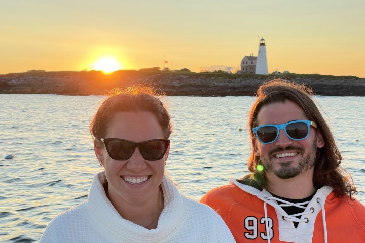 Two people smiling with sunglasses, ocean and lighthouse in the background at sunset.