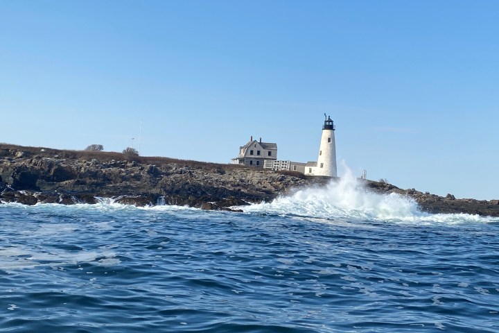 Lighthouse and house on rocky shore with waves crashing, under a clear blue sky.
