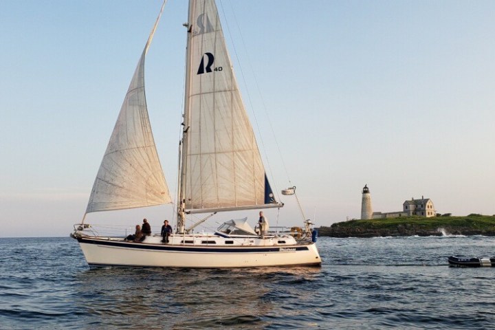 Sailboat on open water with distant lighthouse and building on the horizon under a clear sky.